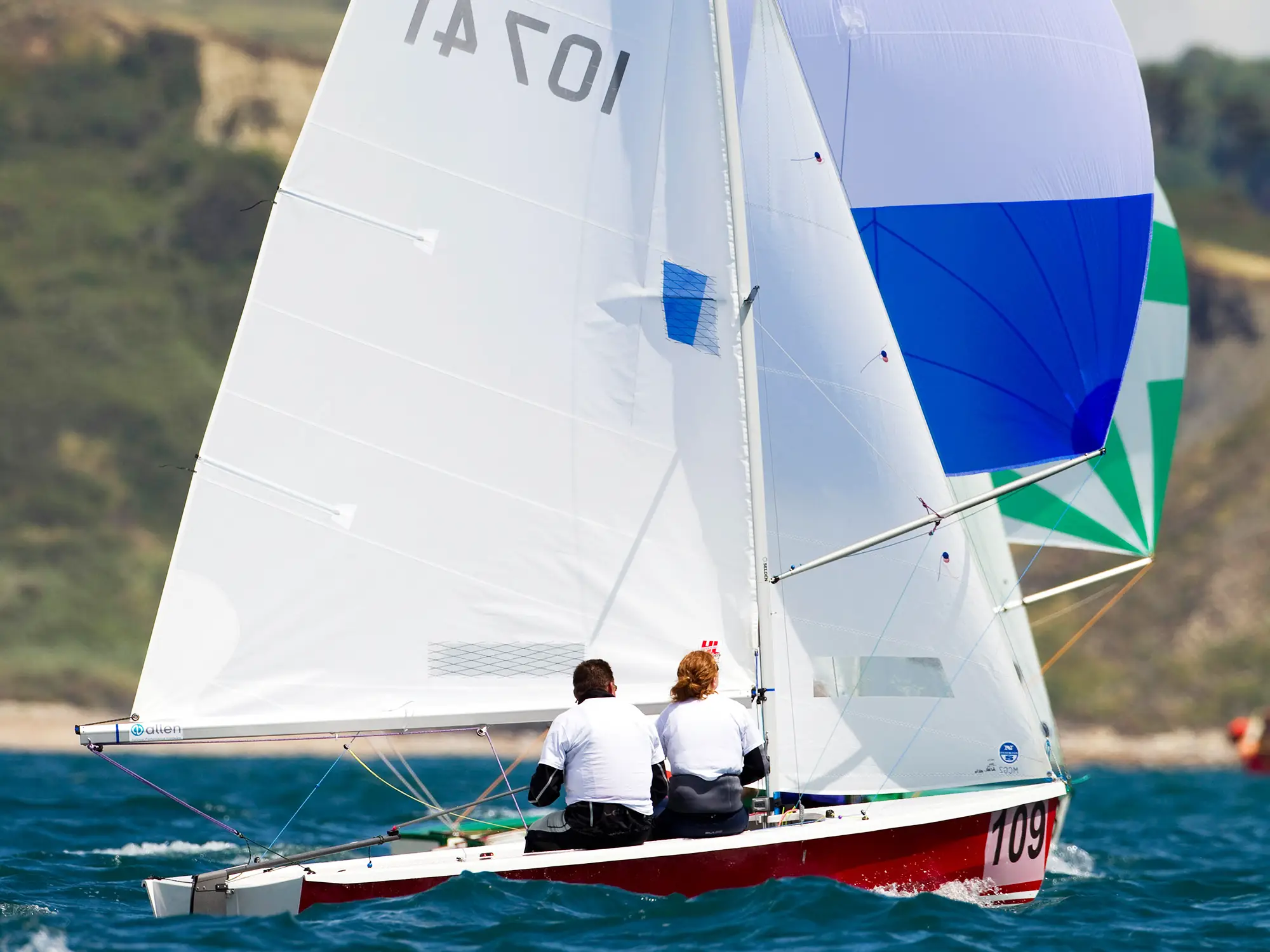 Two-person dinghy racing under spinnaker in open water, crew hiking out on port tack